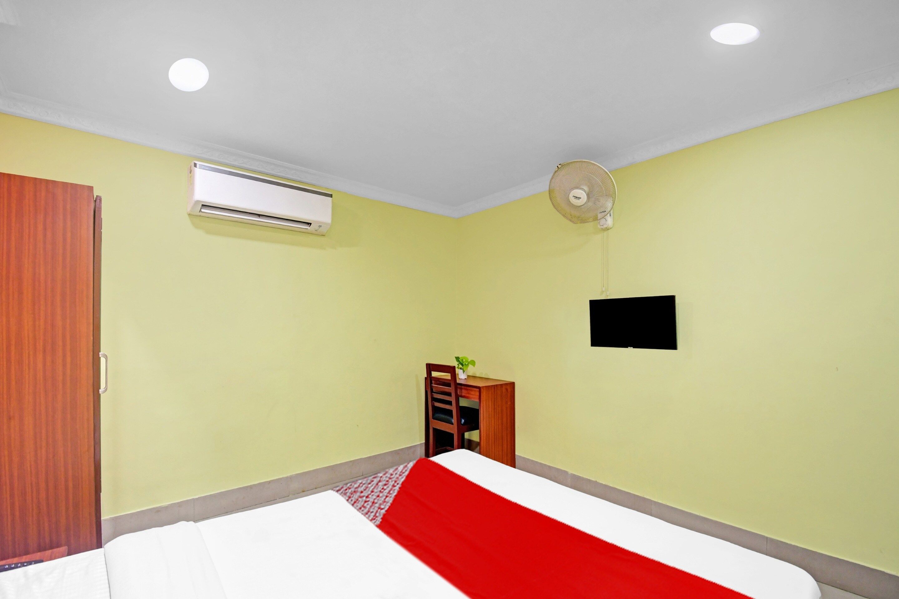Una habitación con una cama, un aire acondicionado, un ventilador de pie, un armario de madera y una mesita con una planta.
