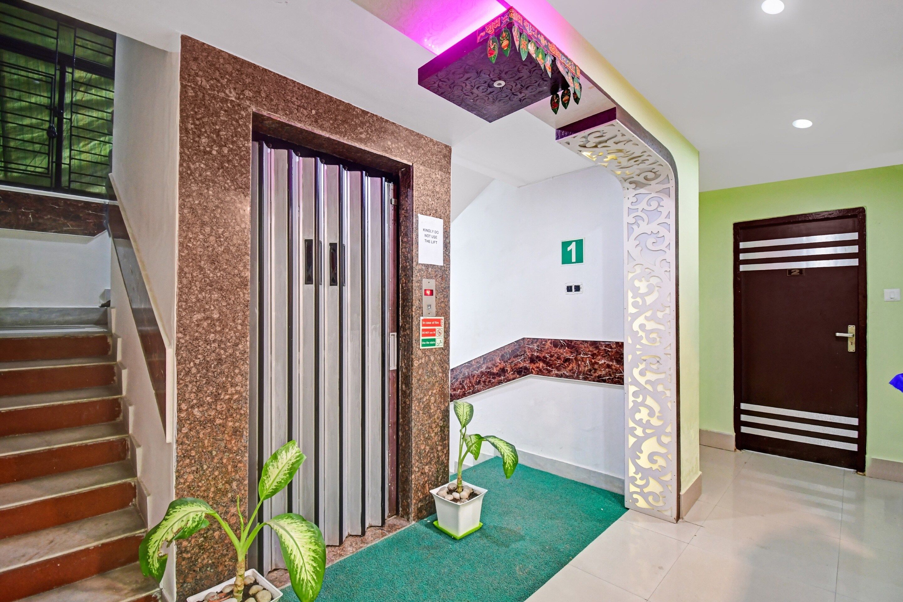 Un vestíbulo de hotel con una escalera, un ascensor, una zona alfombrada de color verde y un techo decorativo.