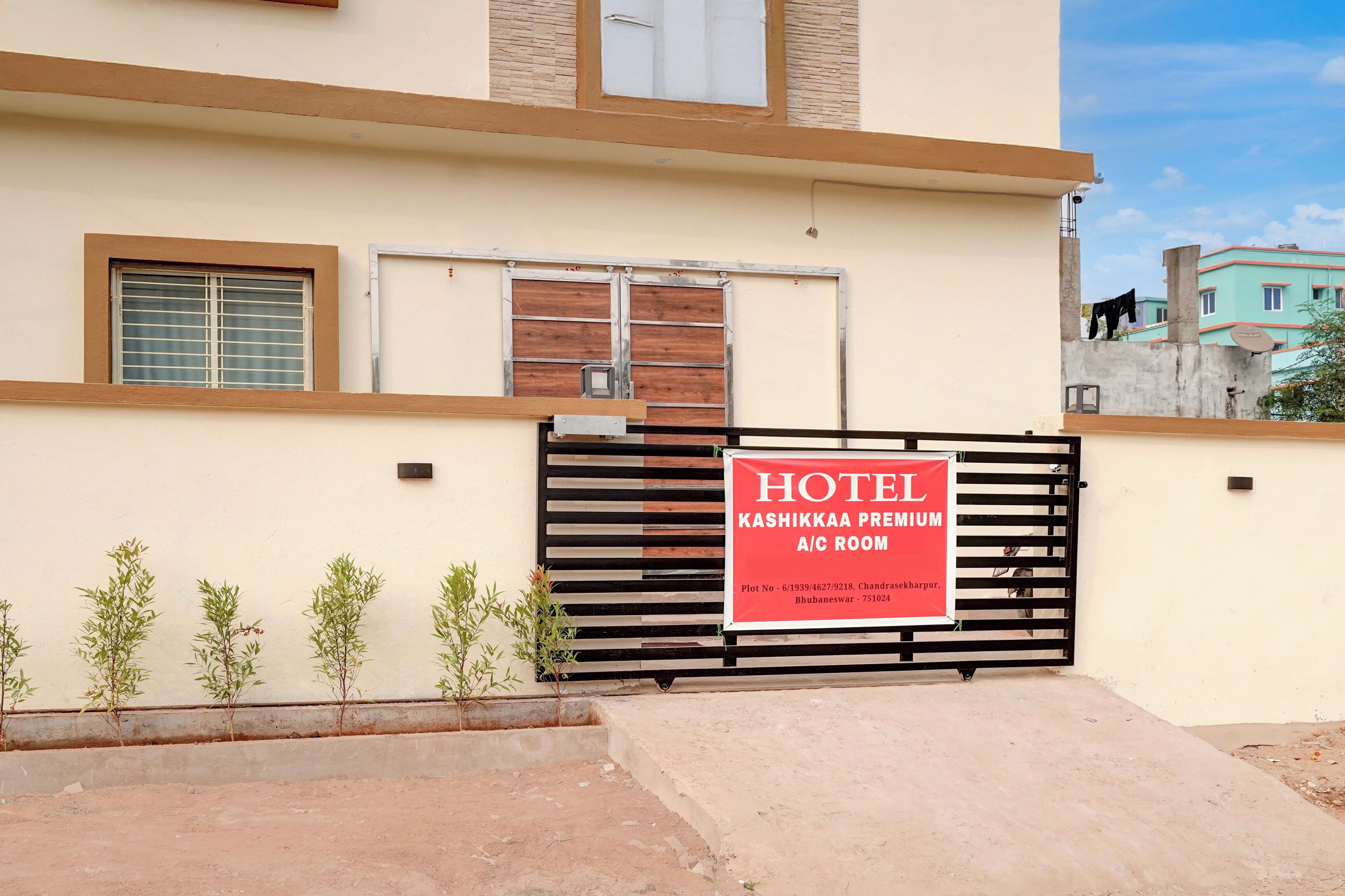 A hotel entrance with a gated driveway, a signboard for 'Kashikkaa Premium A/C Room,' and a view of the surrounding buildings.