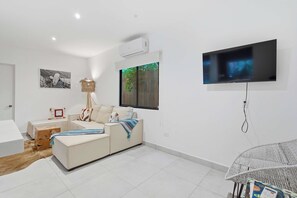 A modern living room with a sofa, a TV mounted on the wall, and a window with a view of greenery. - Private Villa with pool and BBQ (Potrero)