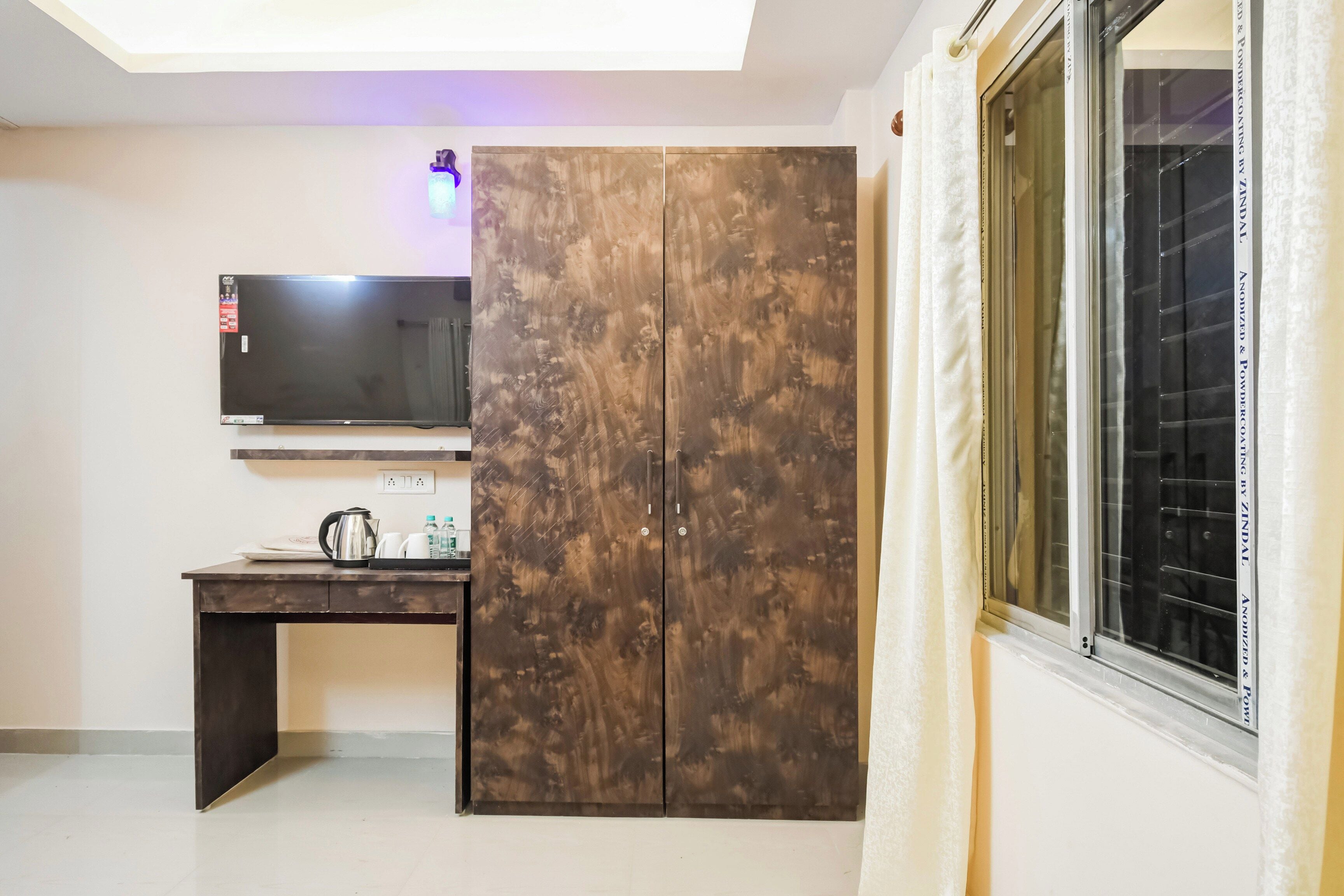A room with a TV mounted on the wall, a wooden wardrobe, a desk with a kettle, and a window with curtains.