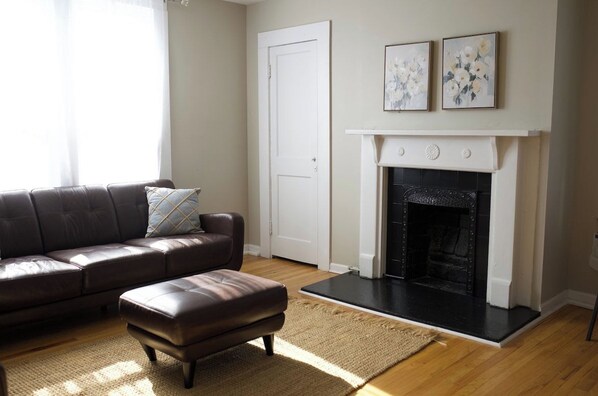 A living room with a leather sofa, a matching ottoman, a fireplace, and two framed artworks on the wall.
