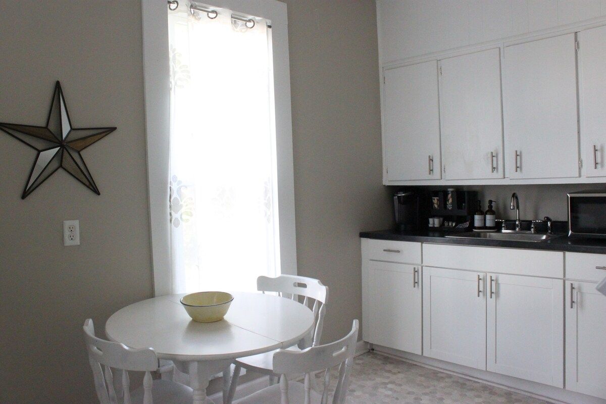 A kitchen with white cabinets, a white table with chairs, a star decoration on the wall, and a window with curtains.