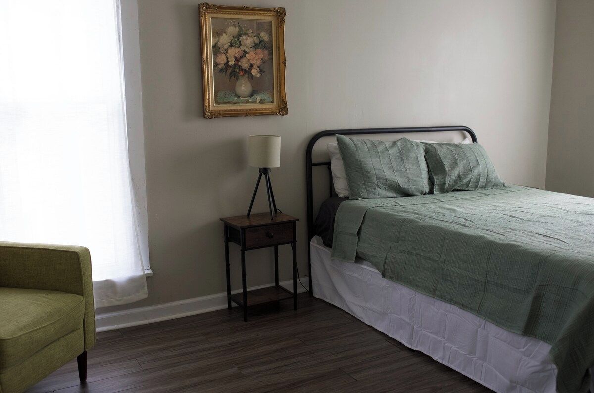 A bedroom with a bed, a nightstand, a lamp, a chair, and a framed picture on the wall.