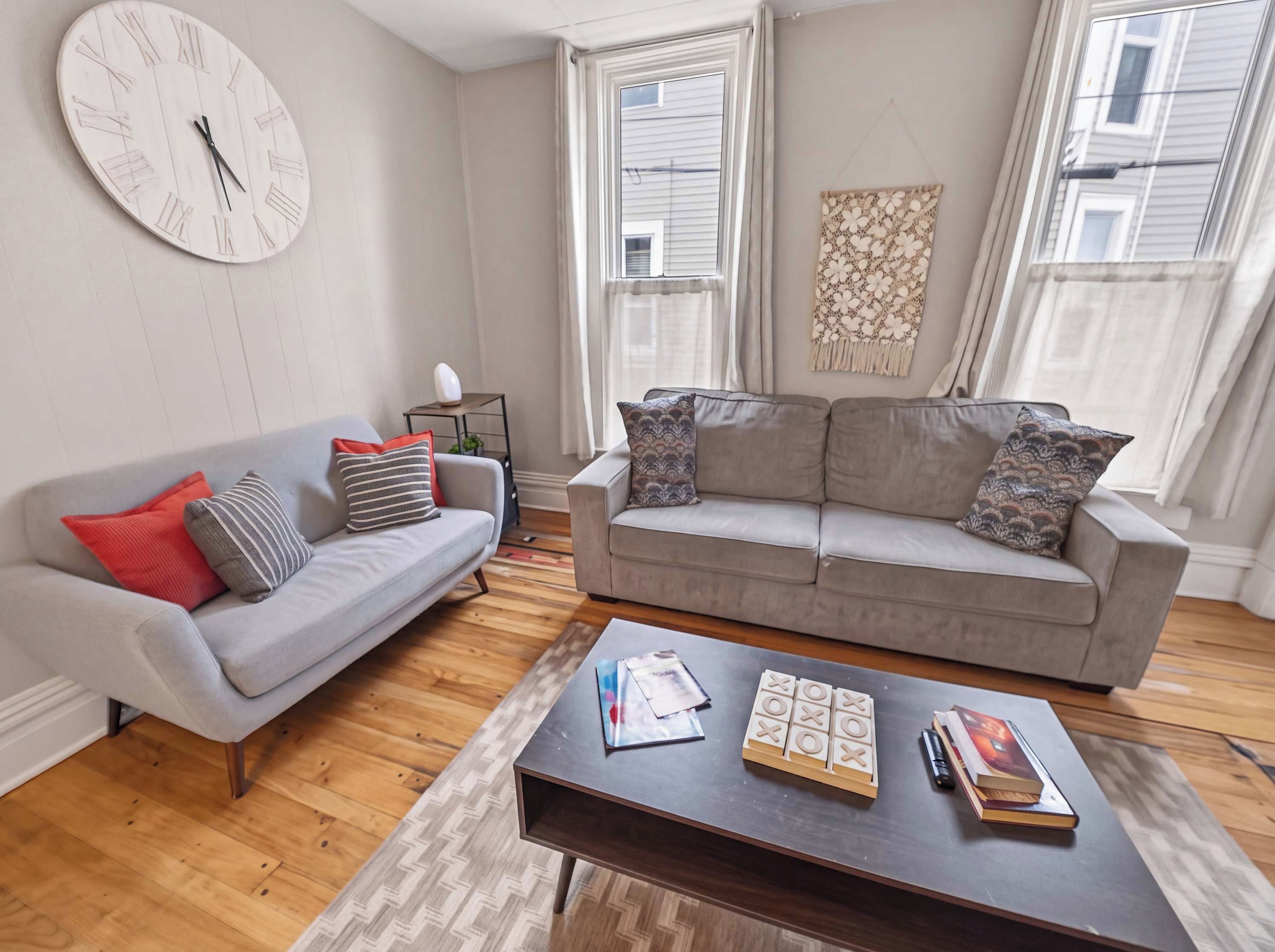 A living room with a grey sofa, a grey armchair, a wooden coffee table with books and a game, and a wall clock.