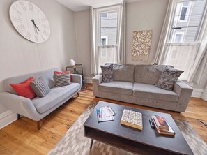 A living room with a grey sofa, a grey armchair, a wooden coffee table with books and a game, and a wall clock.