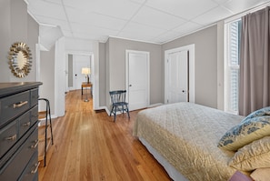 A bedroom with a bed, dresser, chair, and a hallway leading to another room.