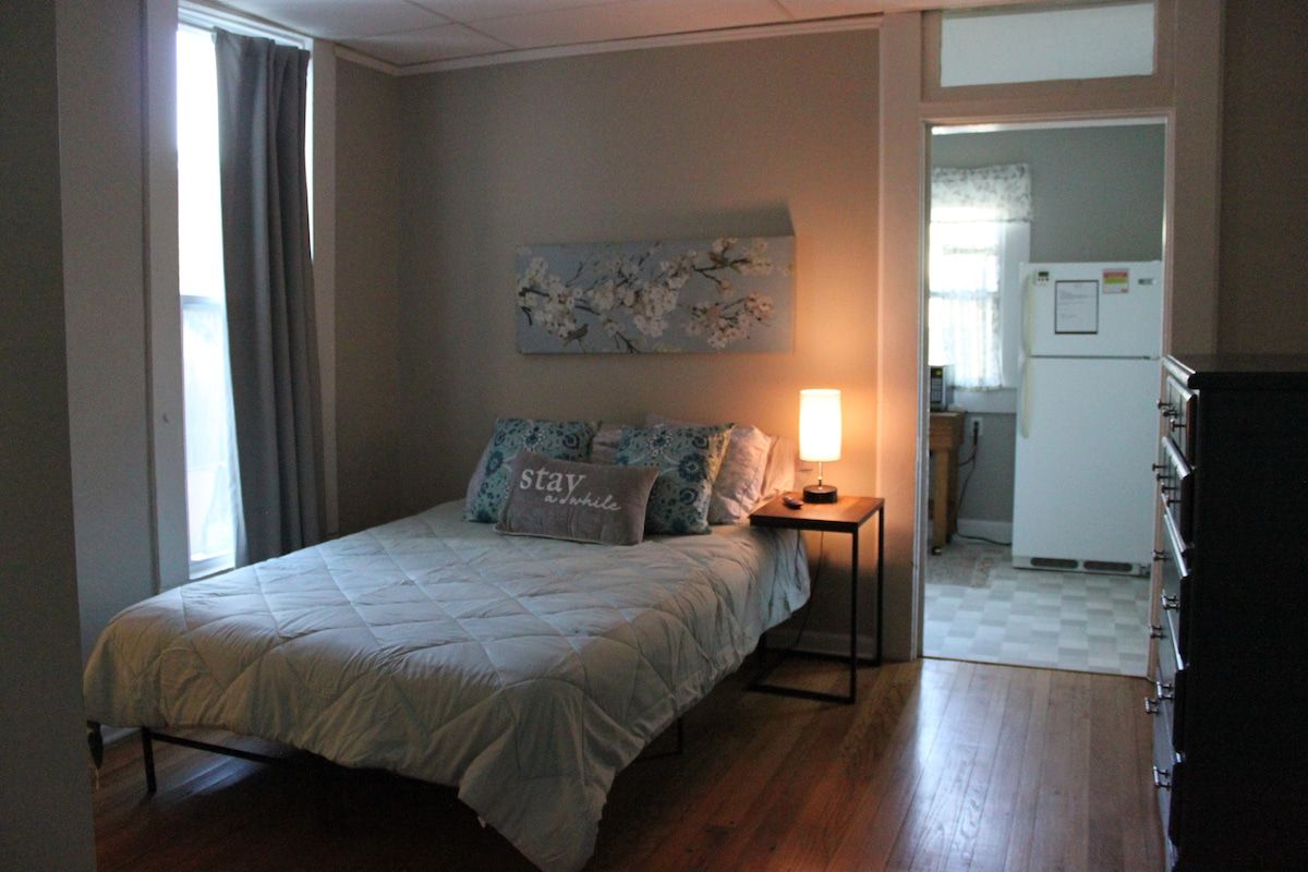 A bedroom with a bed, a nightstand, a wall-mounted lamp, a painting, and a view of a kitchen.