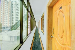 Un pasillo con una puerta de madera a la derecha, una ventana a la izquierda y vistas de edificios altos al exterior.