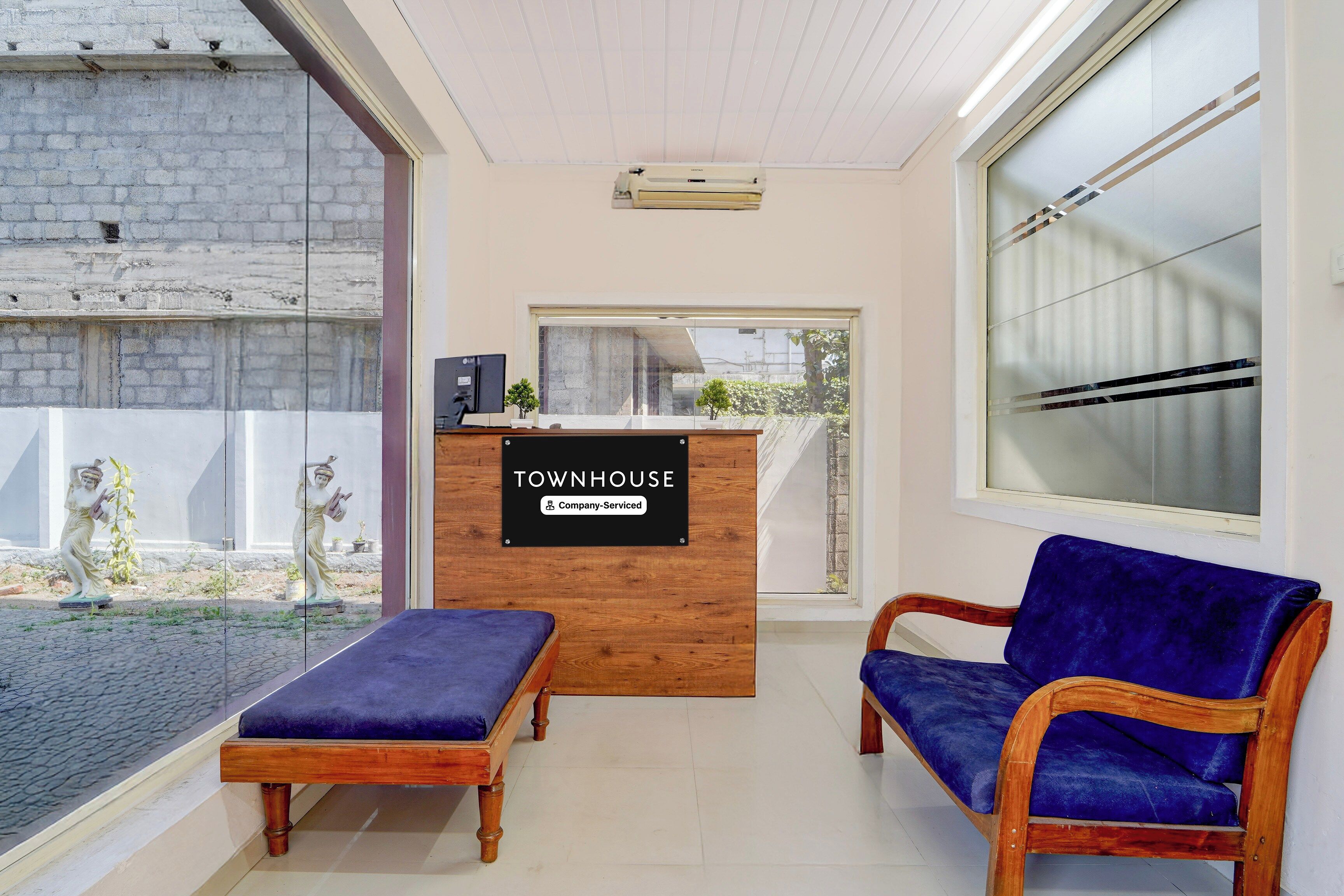 A reception area with a wooden desk, two blue cushioned chairs, a statue outside, and a large window.