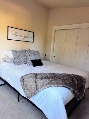 A neatly made bed with a fluffy blanket, a wall with a 'be our guest' sign, and a closed door. - The Oakway - Close to UO Campus + Autzen + Golf! (Eugene)