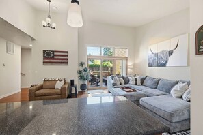 A spacious living room with a large grey sectional sofa, a brown leather couch, and a granite coffee table. There is a large window providing natural light and a view of the outdoors. - The Oakway - Close to UO Campus + Autzen + Golf! (Eugene)