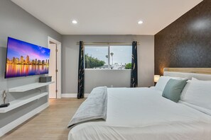 A modern bedroom with a large bed, a flat-screen TV on a wall, a window with curtains, and a view of the cityscape. - Modern 3BR | Walk to Bay • Pet-Friendly • Parking (San Diego)