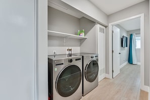 A laundry room with two front-loading washing machines, a shelf with cleaning supplies, and a door leading to another room. - Modern 3BR | Walk to Bay • Pet-Friendly • Parking (San Diego)
