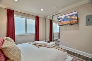 A hotel room with a bed, a TV mounted on the wall, and a large window with red curtains. - Surf City | Rooftop Fire Pit | Walk to Pier (Huntington Beach)