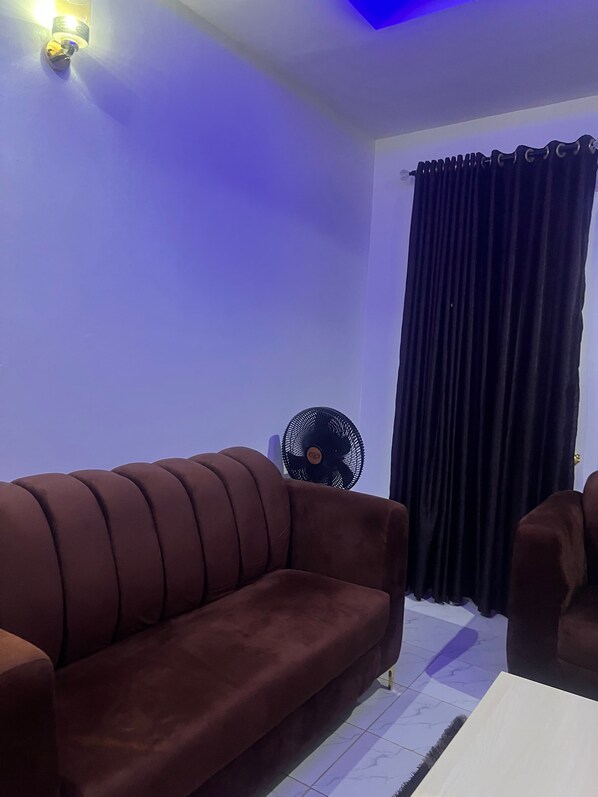 Una habitación con un sofá marrón, un ventilador de pie y una ventana con cortinas oscuras.