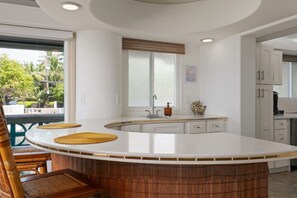 A modern kitchen with a large island, white cabinetry, and a window with a view of greenery. - Sunsets on the Shoreline | Oceanfront Kona Home (Kailua-Kona)