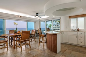 A spacious kitchen with a central island, large windows offering ocean views, and a dining area with wicker furniture. - Sunsets on the Shoreline | Oceanfront Kona Home (Kailua-Kona)