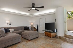 Una sala de estar con sofá, reposapiés, televisor y ventilador de techo.