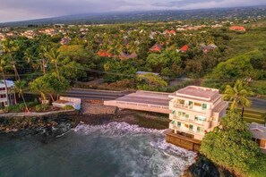 A coastal building with a long structure adjacent to the sea, surrounded by greenery and other buildings. - Sunsets on the Shoreline | Oceanfront Kona Home (Kailua-Kona)