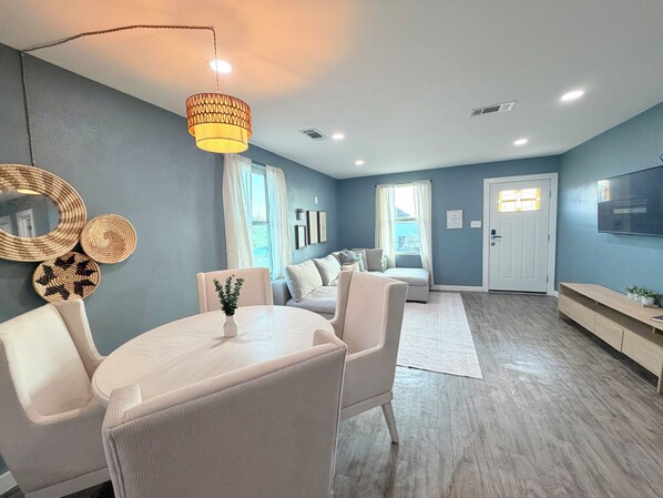 A modern living room with a dining area, a sofa set, a TV mounted on the wall, and a large window with curtains.