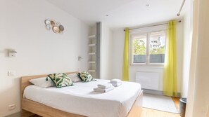 Quarto com cama, almofadas, mesa de cabeceira, janela com cortinas e luminária na parede.
