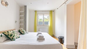 A bedroom with a bed, pillows, a window with curtains, a radiator, and a small stool. - L’Île Verte – Auditorium – Exhibition Centre (Dijon)