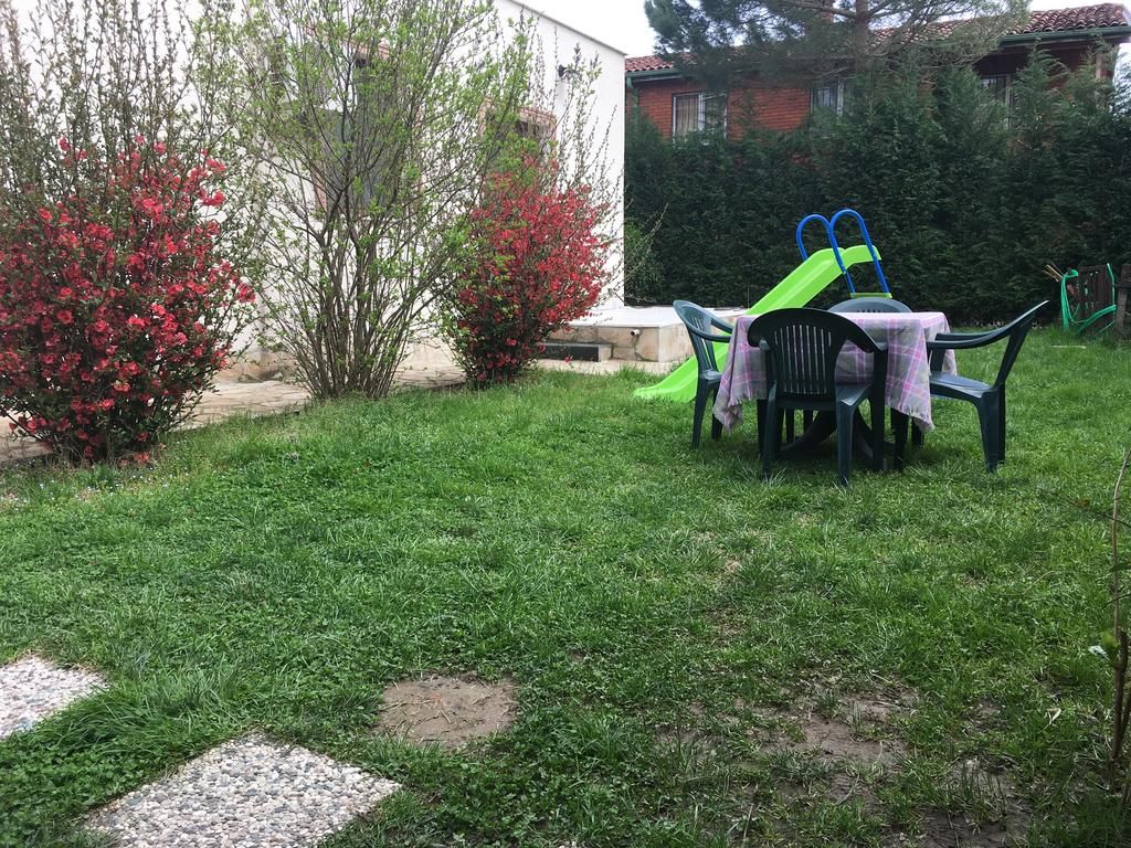 A grassy outdoor area with a table and chairs, a green slide, and red flowering bushes.