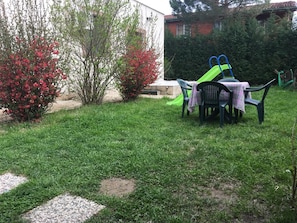 Un espacio al aire libre con césped, una mesa y sillas, un tobogán verde y arbustos rojos con flores.