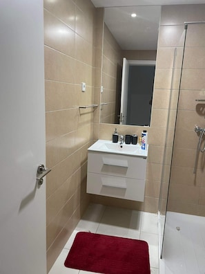 A modern bathroom with a glass shower enclosure, a white sink with a mirror above it, and a red bath mat on the floor. - Adorable 2-bedroom apartment in vibrant Mindelo perfect for relaxing stay (Mindelo)