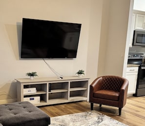 A living room with a flat-screen TV mounted on the wall, a wooden entertainment unit, a brown leather armchair, and a grey tufted ottoman. - Beautiful House in Sebring! Sleeps 8 (Sebring)