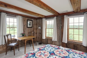 Quarto com paredes e teto de madeira, uma cama com colcha, uma mesa com cadeiras e janelas com cortinas.