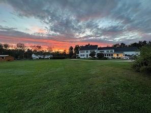 Uma casa grande e branca com varanda, cercada por um campo gramado e árvores.
