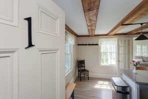 Una habitación con techo de madera, paredes blancas, un banco, una silla, un ventanal y una zona de cocina con lavaplatos y encimera.