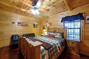 A log cabin-style bedroom with a bed, a wooden nightstand, a desk, a chair, a ceiling fan, and a window with blinds. - Southern Grace: Hot Tub, Fire Pit & Walkable Location (Broken Bow)