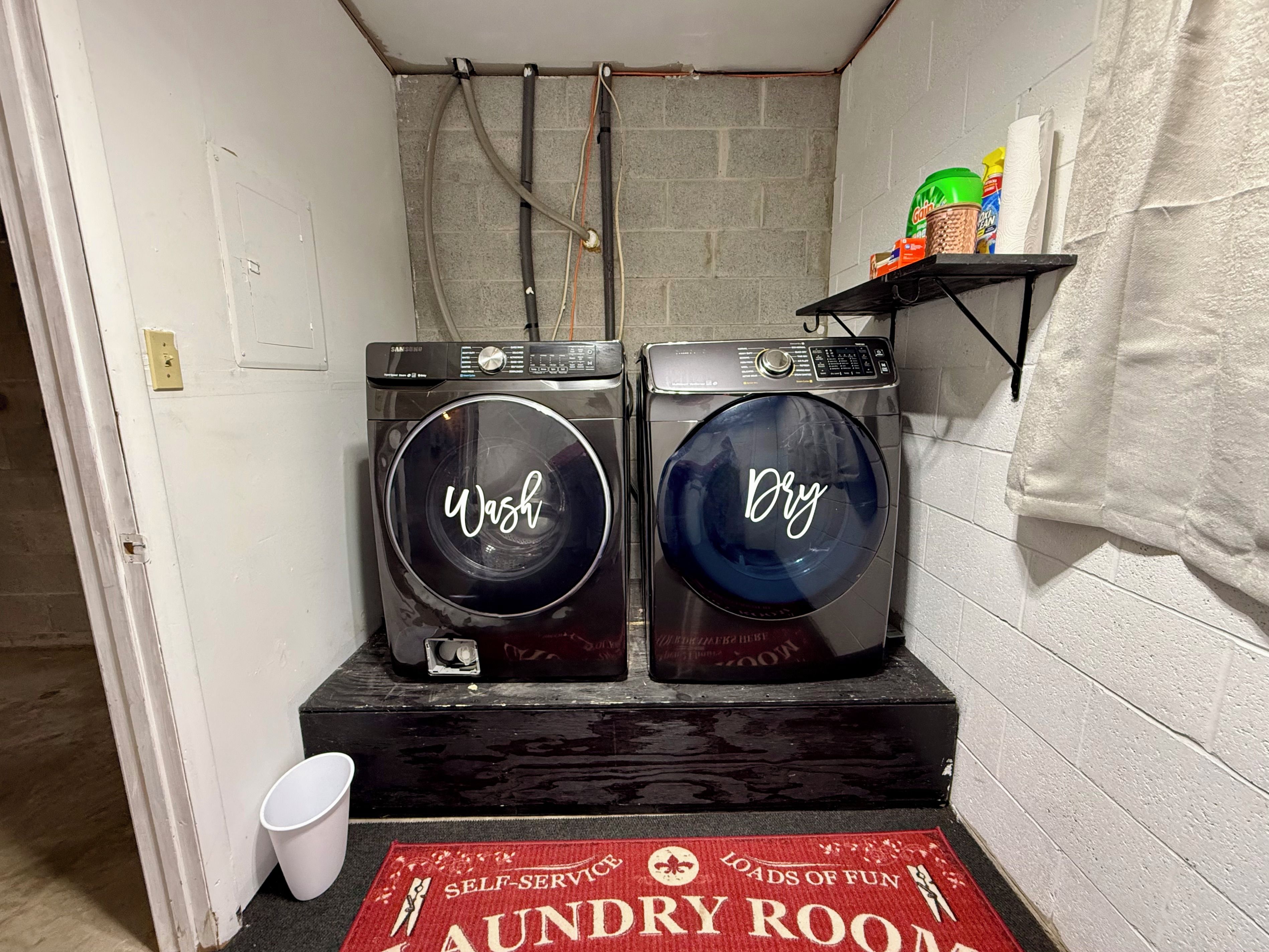 A laundry room with two front-loading washing machines, a red mat with 'Laundry Room' and 'Self-Service' text, and a shelf with cleaning supplies.
