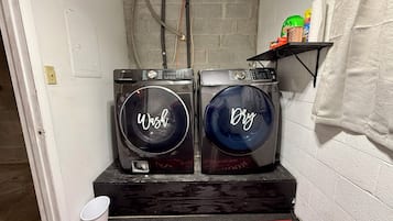 A laundry room with two front-loading washing machines, a red mat with 'Laundry Room' and 'Self-Service' text, and a shelf with cleaning supplies.