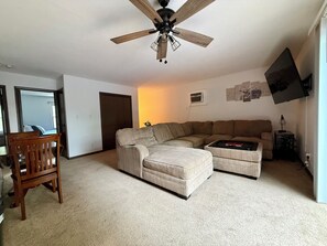 Ein Wohnzimmer mit einem Ecksofa, einem Holzstuhl, einem Deckenventilator und einem an der Wand befestigten Flachbildfernseher.
