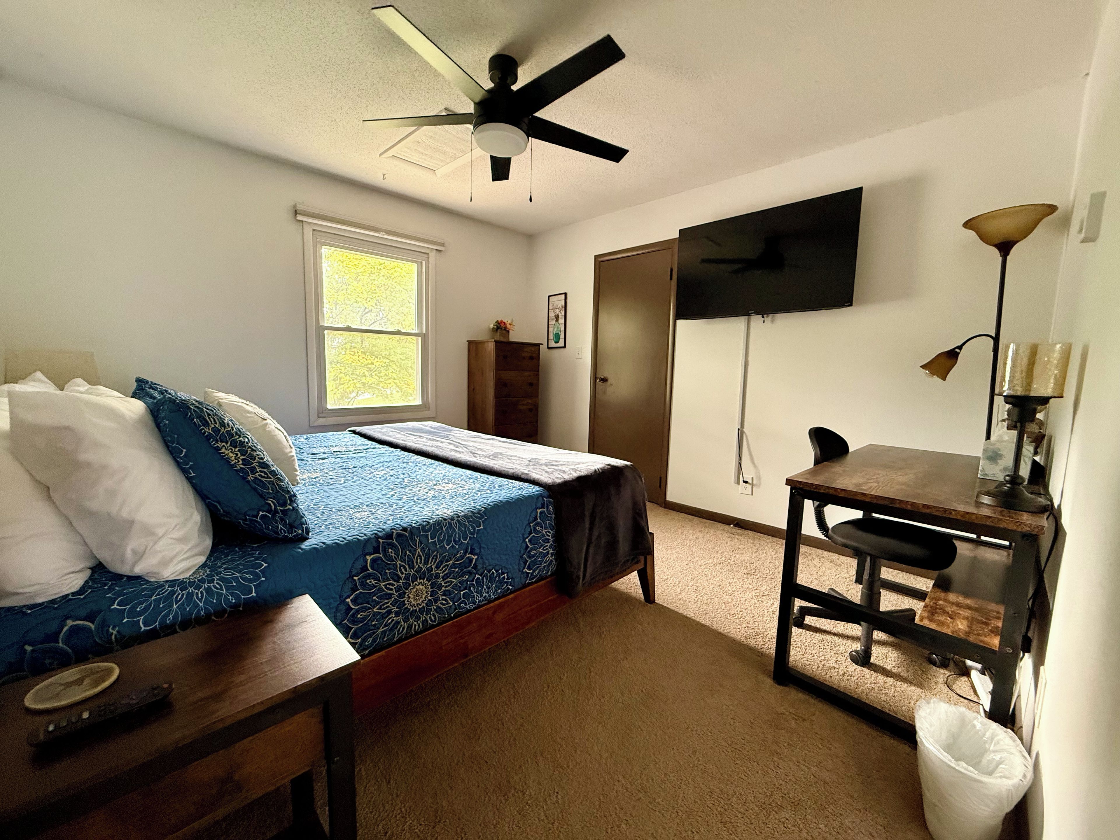 Un dormitorio con cama, escritorio, silla, ventilador de techo, ventana y televisor montado en la pared.