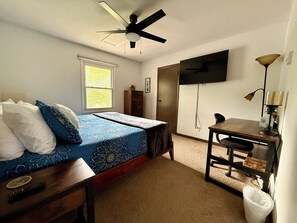 A bedroom with a bed, a desk, a chair, a ceiling fan, a window, and a TV mounted on the wall.