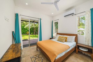 A bedroom with a large bed, a ceiling fan, an air conditioning unit, a bedside table with a lamp, and a view of a garden through a sliding door. - Private Pool Villa • Gated • Near Playa Negra (Provincia de Guanacaste)