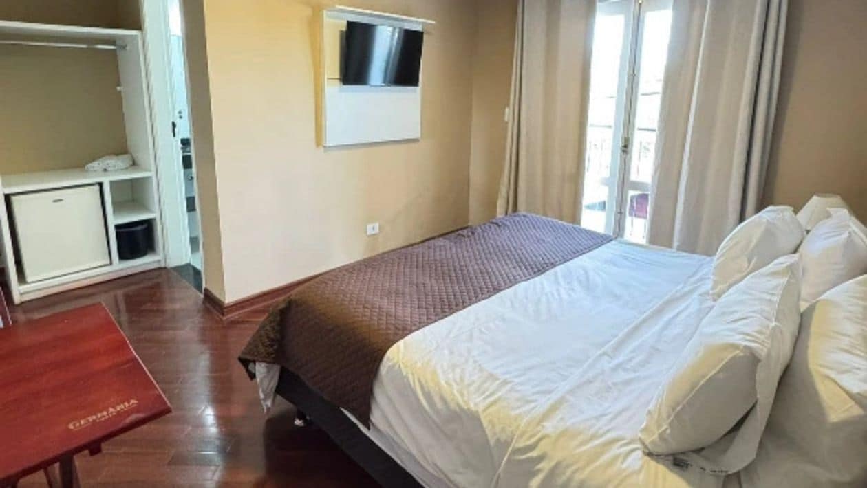 Habitación de hotel con cama, televisor montado en la pared, armario y una pequeña mesa.