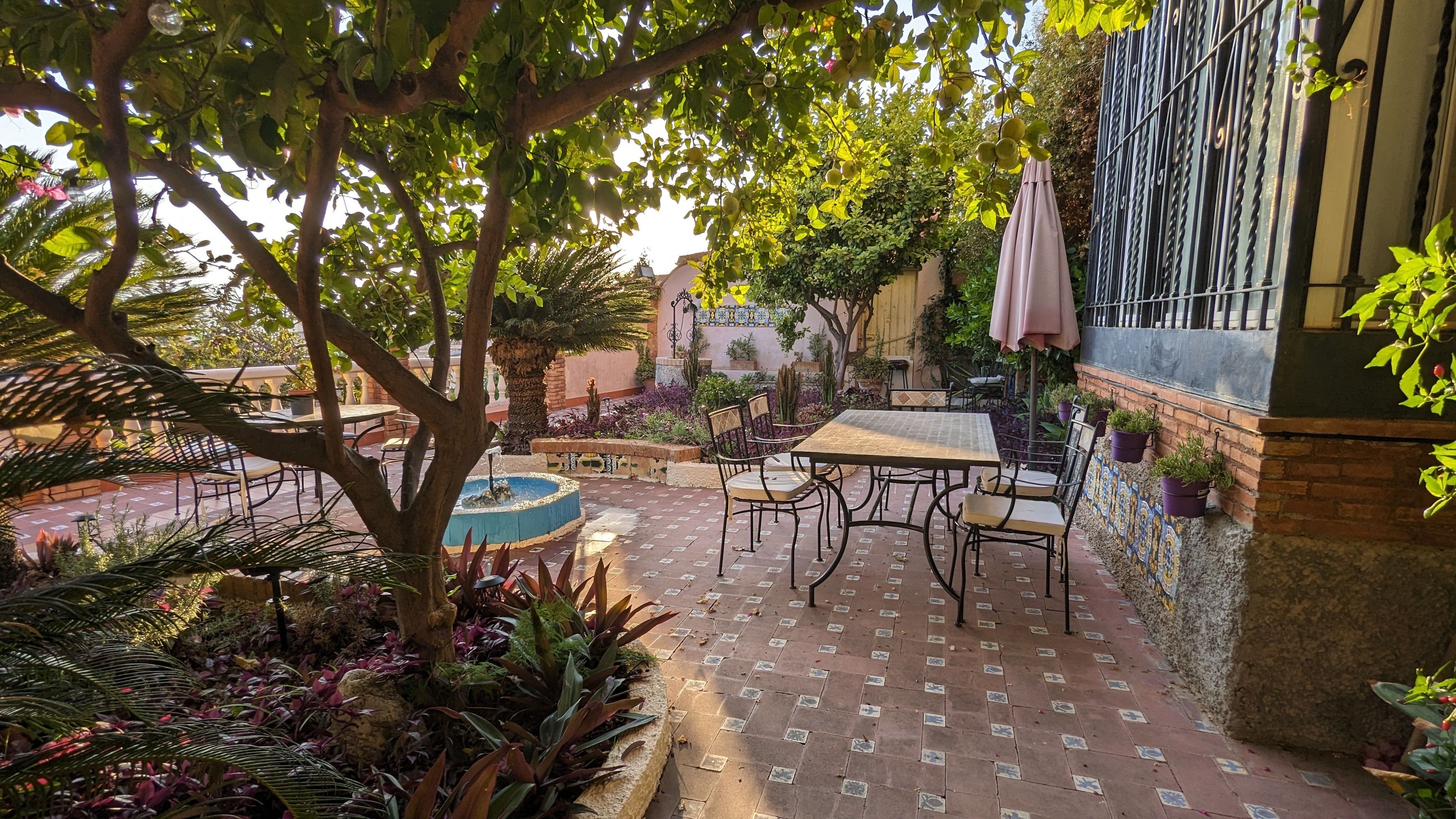 An outdoor patio with a table and chairs, surrounded by potted plants and a fountain.
