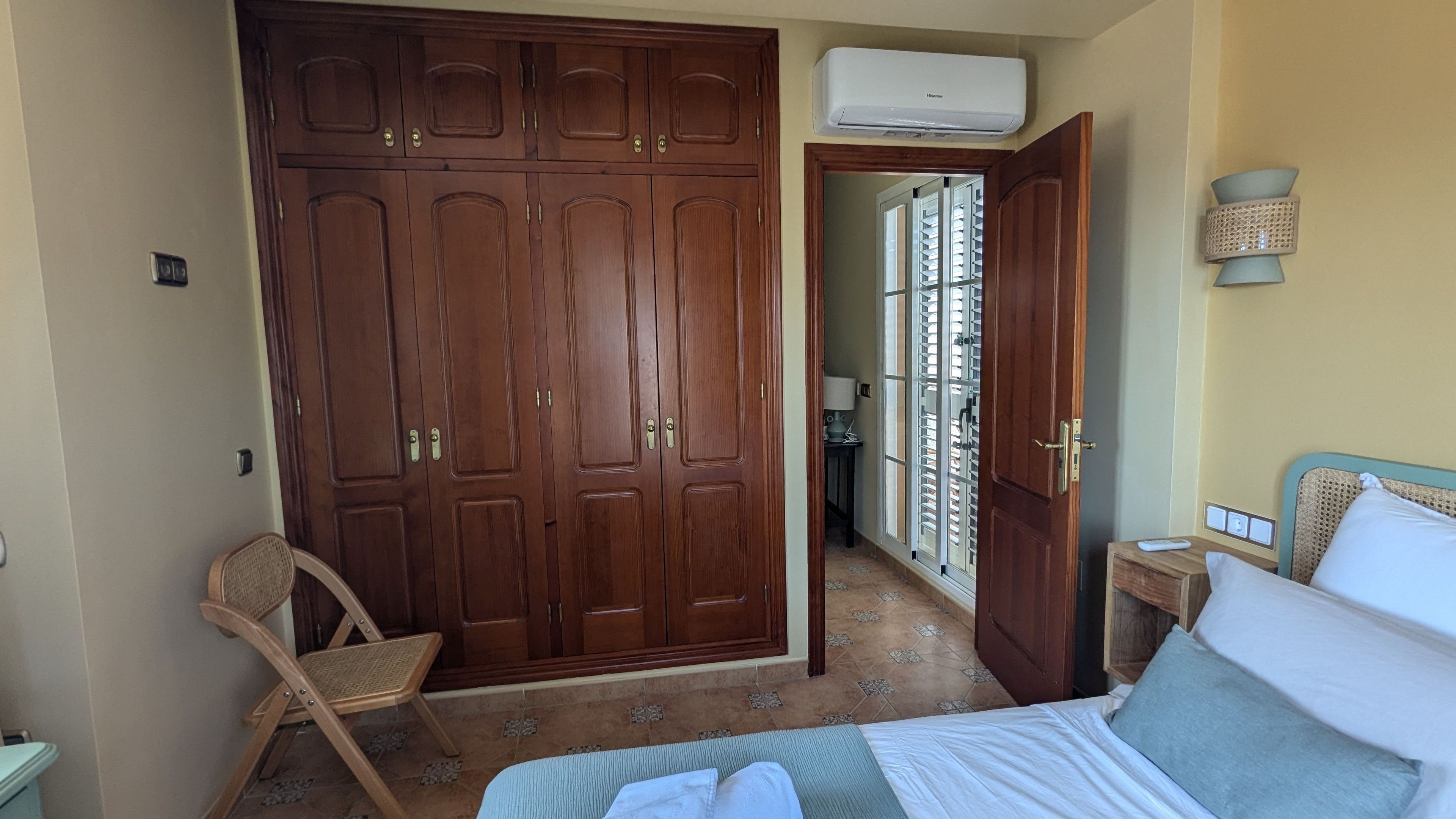 A bedroom with a large wooden wardrobe, a bed with pillows, a chair, and an air conditioning unit.