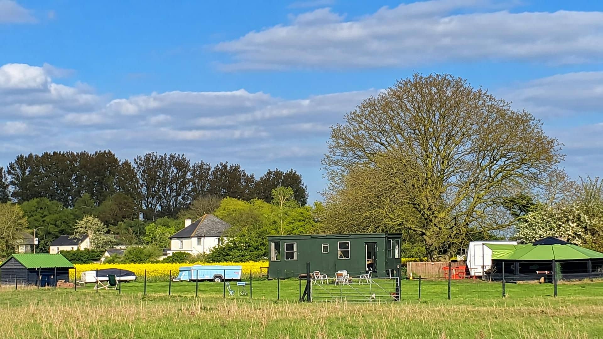 Paisaje rural con una casa verde, una caravana azul y un árbol grande.