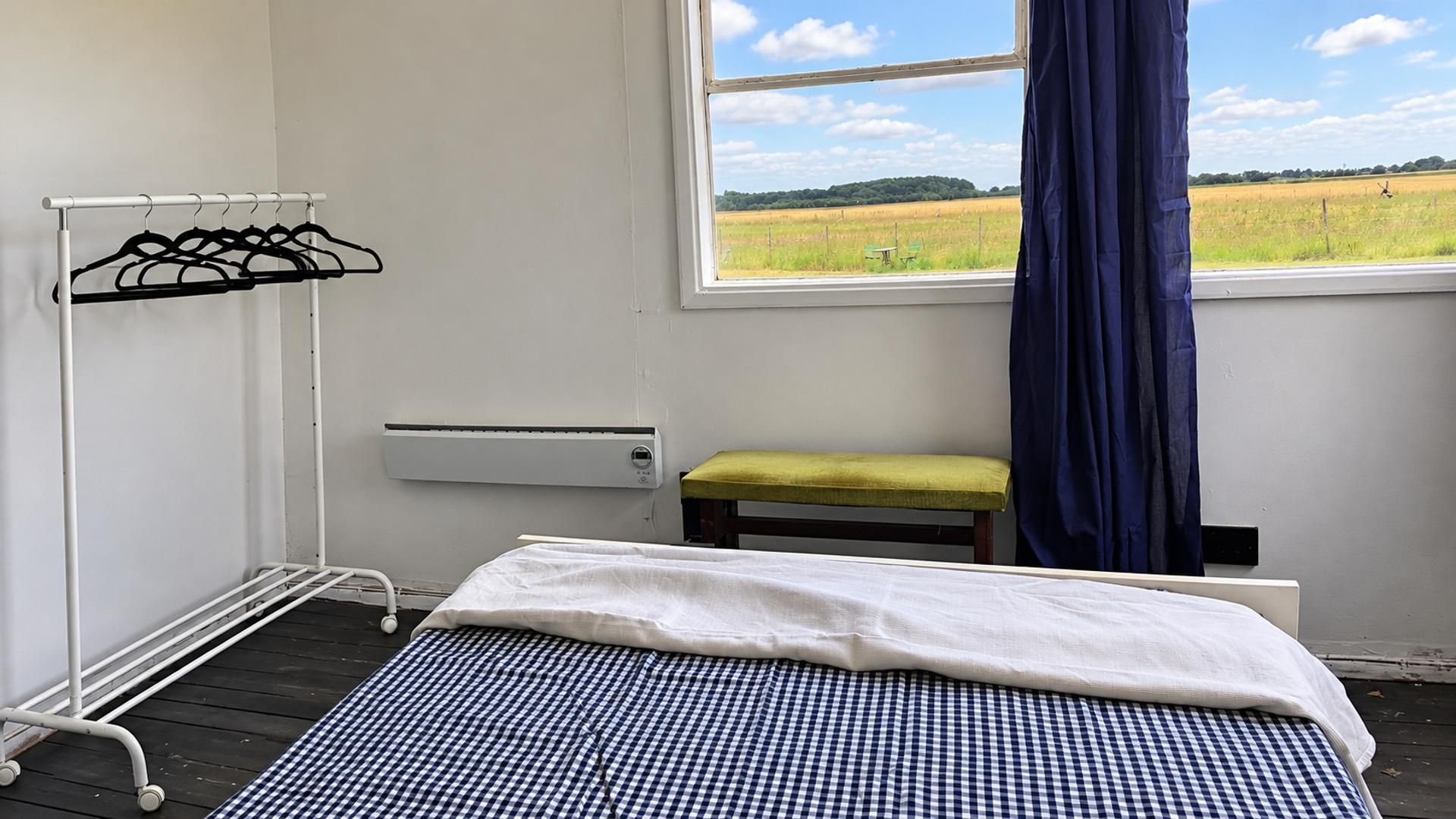 Una stanza con un letto, una panca, un termosifone, un appendiabiti e una finestra vista campo.