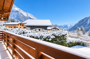 A wooden balcony with snow-covered plants, a mountain backdrop, and a building with a snow-covered roof. - New Listing (Chamonix-Mont-Blanc)