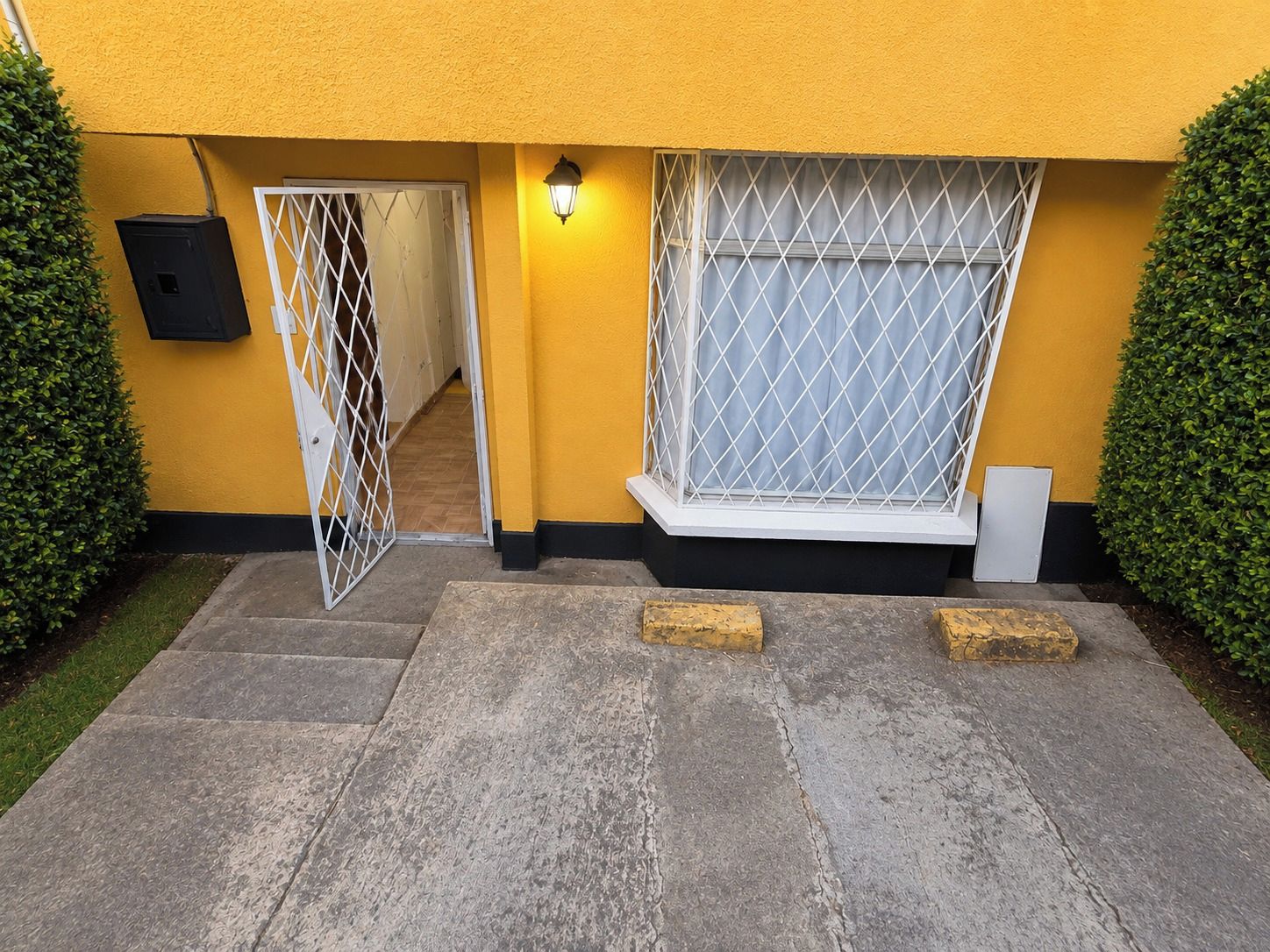 Un edificio amarillo con entrada protegida por una reja, un accesorio de iluminación sobre la puerta y una ventana con celosía de metal.