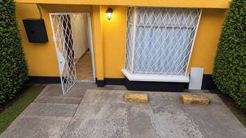 Un edificio amarillo con entrada protegida por una reja, un accesorio de iluminación sobre la puerta y una ventana con celosía de metal.
