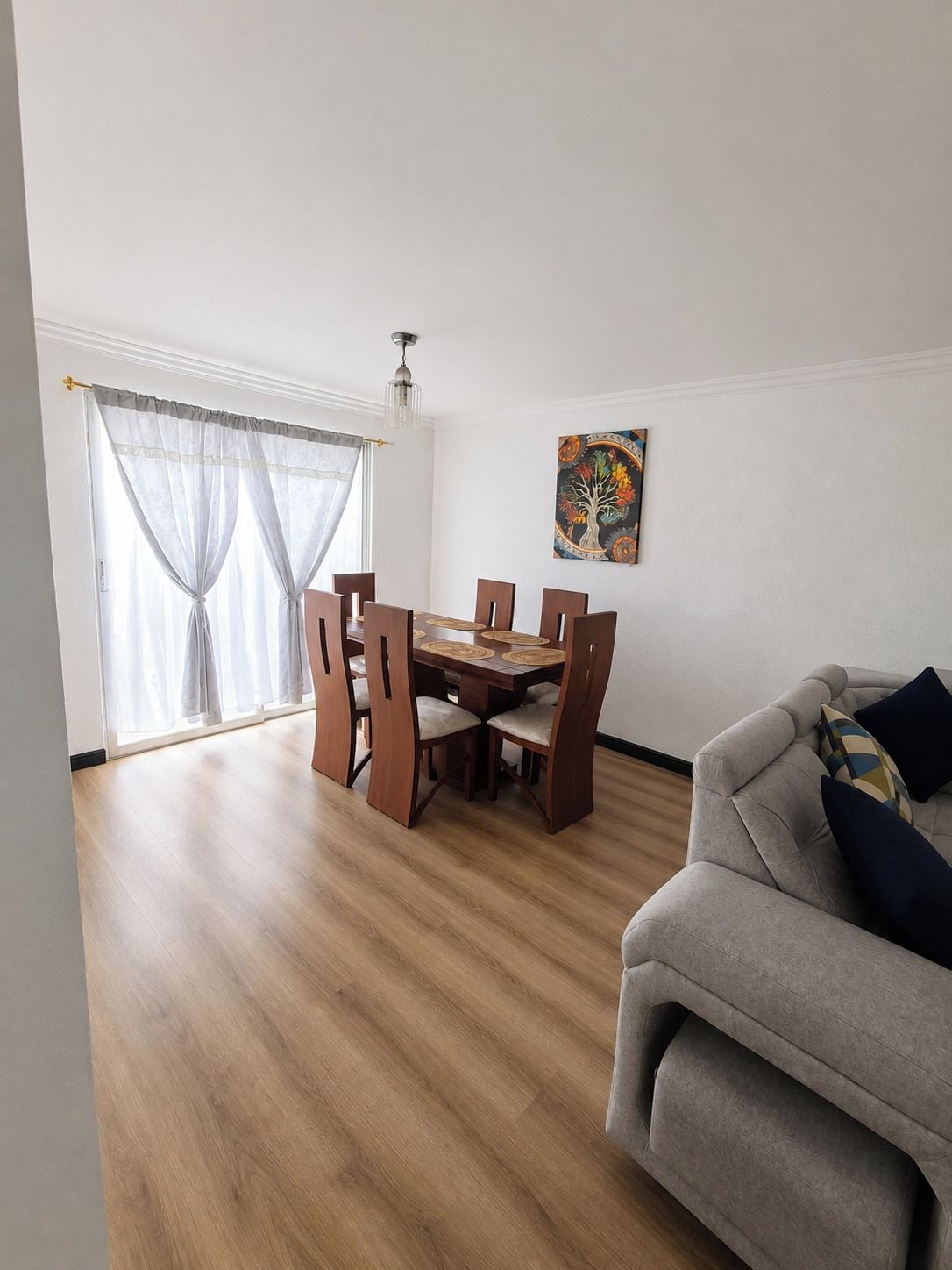 Een eetgedeelte met een houten tafel en stoelen, een bank en een raam met dunne gordijnen.
